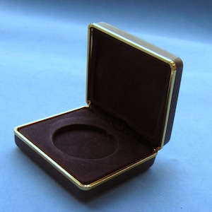 Pode incluir: Uma caixa forrada de veludo marrom escuro com acabamento dourado. A caixa está aberta e a tampa está levantada.