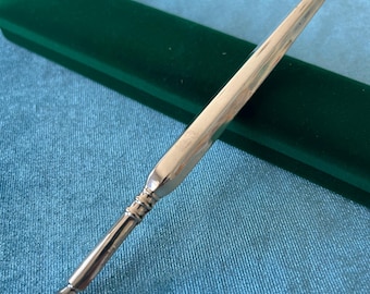 May include: A metallic dip pen with a smooth, tapered handle and a pointed nib. The pen features a polished, reflective surface with a decorative ribbed section near the nib. The design is slender and elongated, showcasing a clean, minimalist aesthetic.