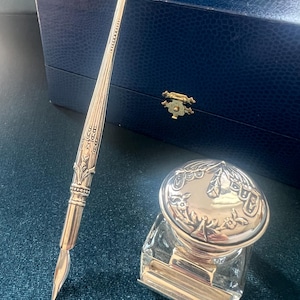 May include: A silver-colored dip pen and inkwell set. The pen has an ornate design and a pointed nib. The inkwell is glass with a silver lid featuring a floral pattern. A blue box is in the background.