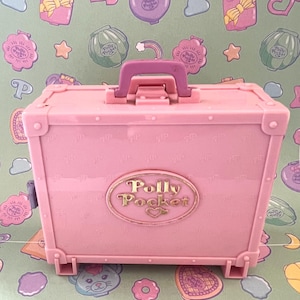 Op de afbeelding: Een roze Polly Pocket koffer met een paarse handgreep. De koffer heeft een gouden ovaal embleem met de woorden "Polly Pocket" en een hart. De achtergrond is een groen oppervlak met patronen en verschillende Polly Pocket ontwerpen.