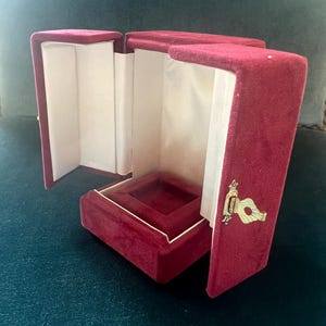 Pode incluir: Uma caixa de joias de veludo bordô com uma tampa articulada, aberta para revelar um interior de cetim branco. A caixa tem formato quadrado com um pequeno compartimento interno. Um fecho dourado prende a tampa.