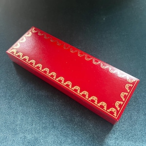 Puede incluir: Una caja rectangular roja con adornos dorados. La caja está cerrada y tiene un acabado brillante. El adorno dorado presenta un patrón ornamental repetido alrededor de los bordes.