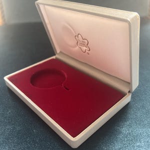 Pode incluir: Uma caixa forrada de veludo branco com um interior de veludo vermelho e um único recorte circular para uma moeda ou medalha. A caixa tem um pequeno logotipo de flor no interior da tampa que diz "All The Good Things In Life Are Free".