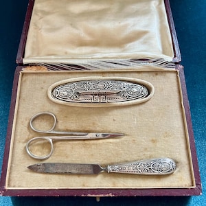 Peut inclure: Un ensemble de manucure en argent dans un coffret doublé de velours rouge. L'ensemble comprend une lime à ongles, des ciseaux et un petit plateau en argent avec un motif orné.