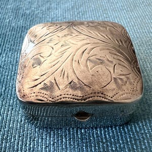 以下が含まれることがあります： 光沢のある仕上げの正方形の金属製小物入れ。蓋には、渦巻く葉とクロスハッチのディテールが施された複雑な手彫りの花柄が描かれています。箱の前面の端には、中央に小さな留め金が見えます。