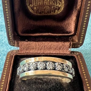 Op de afbeelding: Een zilver- en goudkleurige ring met een centrale band van geoxideerd zilver met een herhalend bloem- of tandwielpatroon, geflankeerd door twee gladde, gepolijste goudkleurige banden, gepresenteerd in een bruin met fluweel bekleed juwelendoosje.