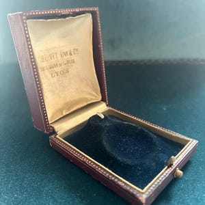 Pode incluir: Caixa de joias antiga aberta, com exterior em couro bordô e interior em cetim dourado. O interior da caixa é forrado com veludo azul escuro. O texto "CHEVET AINE & CO. FOURNISSEURS DE LA VILLE LYON" é visível.