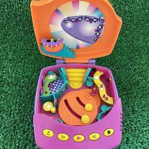 Può includere: Un giocattolo di plastica rosa e arancione con un set di gioco all'interno. Il set di gioco presenta un palco con uno sfondo viola, una scala e una stanza verde e gialla. Il giocattolo ha dei pulsanti sul davanti che dicono "Play", "<<", ">>", e "Stop".