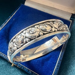 Puede incluir: Una pulsera de plata con diseño floral. La pulsera presenta grabados en relieve detallados de rosas y otras flores. La pulsera se muestra abierta dentro de una caja de joyería azul.