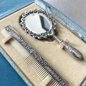 Könnte beinhalten: Ein silberfarbenes Toilettenset bestehend aus einem ovalen Handspiegel und einem rechteckigen Kamm. Der Spiegel hat einen floral geprägten Rand und Griff. Der Kamm verfügt über einen verzierten silberfarbenen Rücken mit Rankenmuster und weiße Zinken.