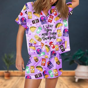 May include: A lilac-colored pajama set featuring a repeating pattern of Taco Bell-themed graphics, including tacos, drinks, and the Taco Bell logo. The shirt has the text "I Love You More Than Taco Bell". The set includes a short-sleeved top and matching shorts.
