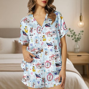 Op de afbeelding: Lichtblauwe pyjamaset met een patroon van Disney-figuren, waaronder Mickey Mouse, Minnie Mouse en Donald Duck. De top met korte mouwen en de shorts hebben witte biezen en zijn versierd met nautische elementen en strikken.