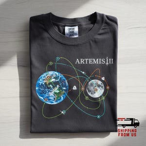 May include: Dark gray t-shirt featuring a graphic of Earth and the Moon with connecting lines and the text "ARTEMIS III". The design includes numbered points and illustrations of spacecraft and buildings.