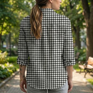 Può includere: Una camicia a quadri bianchi e neri con maniche lunghe, arrotolate sui gomiti. La camicia presenta un classico motivo a quadretti ed è realizzata in un tessuto leggero. La camicia è indossata da una persona.