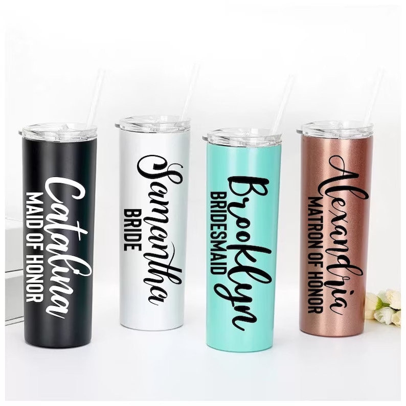 May include: Four personalized stainless steel tumblers with clear straws. The tumblers are black, white, teal, and rose gold. Each tumbler has a name and title in black script: Catalina, Samantha, Brooklyn, and Alexandria.