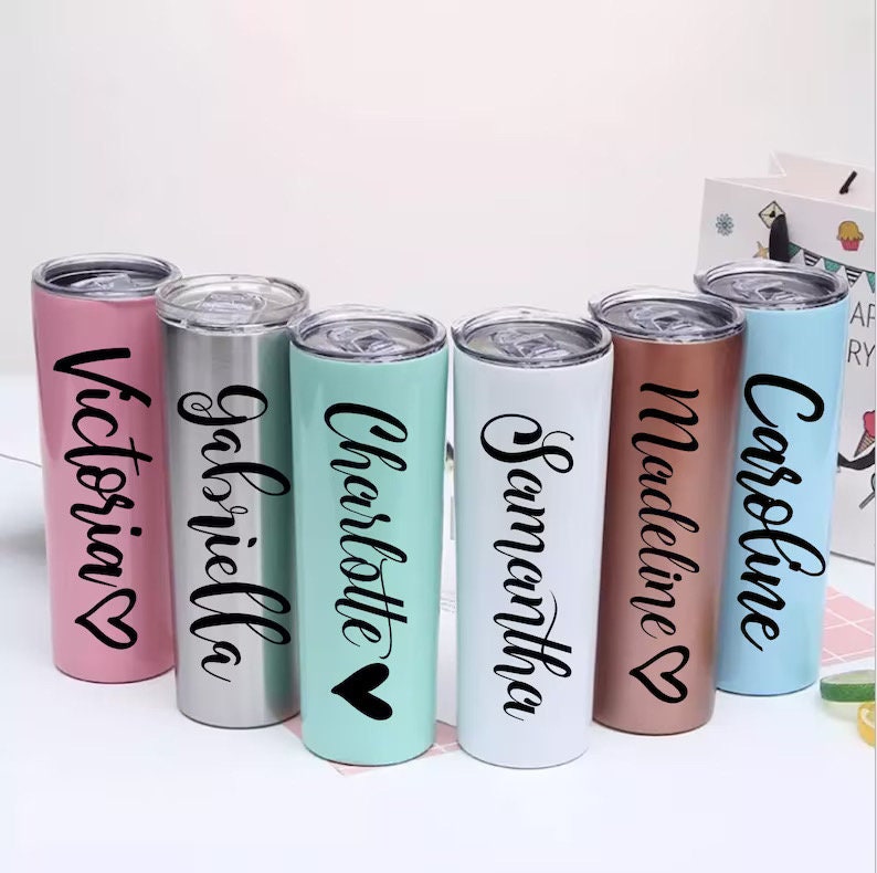 May include: A collection of personalized stainless steel tumblers in various colors, including pink, silver, mint green, white, copper, and light blue. Each tumbler features a name in black script with a heart symbol. The tumblers have clear lids.