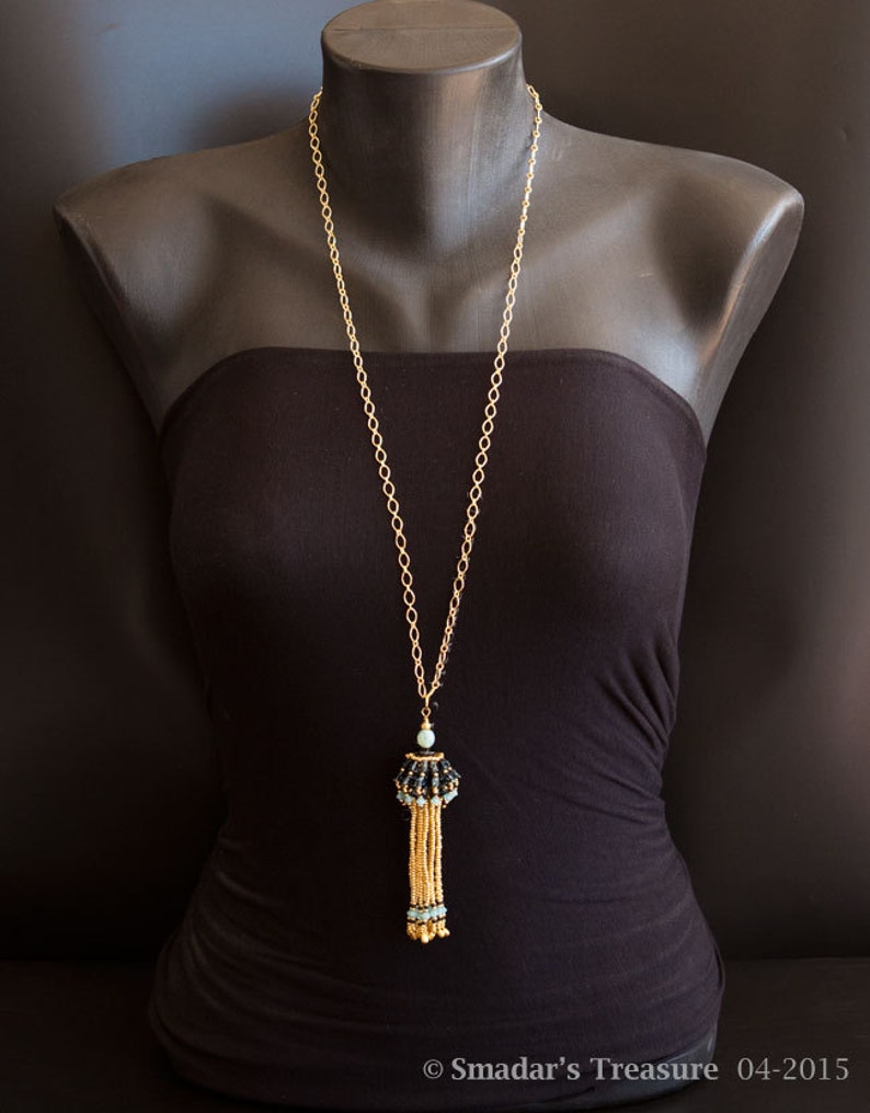 May include: A gold chain necklace with a black and blue beaded tassel pendant. The necklace is displayed on a black mannequin.