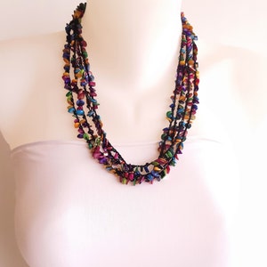 以下が含まれることがあります： カラフルで虹色の平らな木製のビーズでできたマルチストランドネックレス。ビーズは黒いコードに糸で通され、ネックレスは白いマネキンに飾られています。