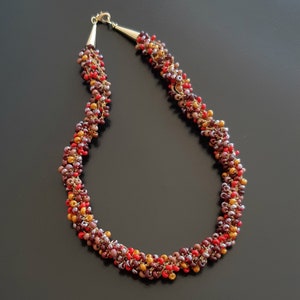 Peut inclure: Un collier de perles brun, rouge et or avec un fermoir en or. Les perles sont petites et regroupées, créant un aspect texturé.