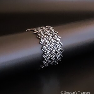 Puede incluir: Un brazalete plateado hecho de pequeñas cuentas cuadradas tejidas juntas en un patrón geométrico.
