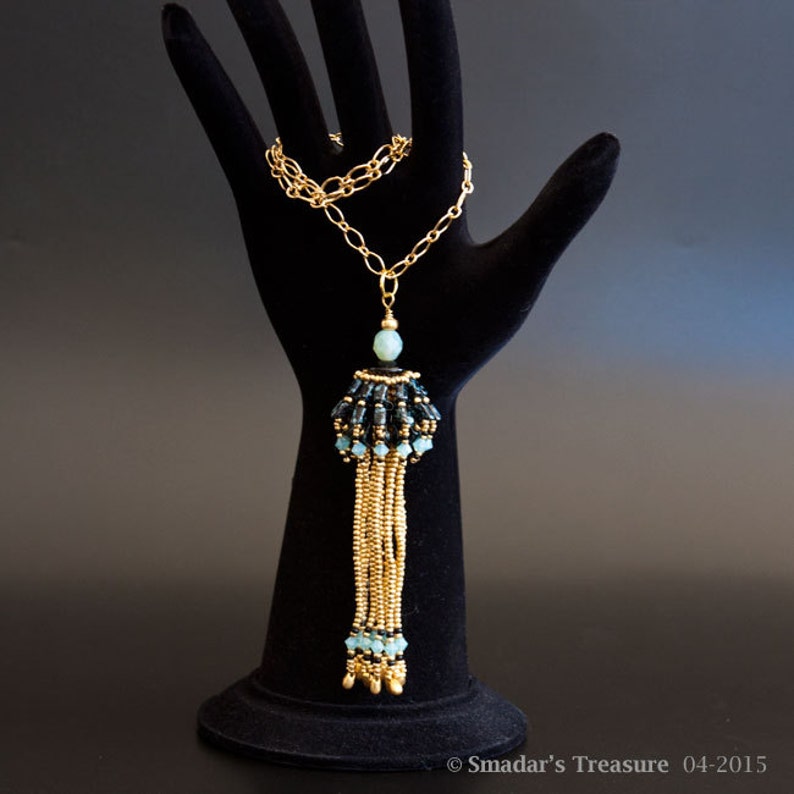 May include: A gold chain necklace with a tassel pendant. The tassel is made of gold beads and black and blue beads. The pendant is topped with a light blue bead.