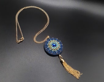 Könnte beinhalten: Eine goldfarbene Halskette mit einem marineblauen Anhänger. Der runde Anhänger ist mit türkis- und goldfarbenen Perlen und einem zentralen blauen Edelstein verziert. Eine goldfarbene Quaste hängt am Anhänger. Die Halskette wird auf einer schwarzen Oberfläche präsentiert.