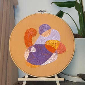 Könnte beinhalten: Ein runder Stickrahmen mit einem beigefarbenen Stoffhintergrund. Der Stoff ist mit abstrakten Formen in Violett-, Orange- und Gelbtönen bestickt.