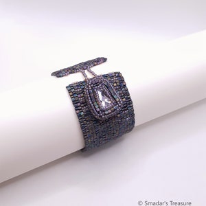 Könnte beinhalten: Ein Armband aus Perlen mit einem großen, klaren Kristall in der Mitte. Das Armband besteht aus kleinen, schillernden Perlen in Violett- und Grautönen.