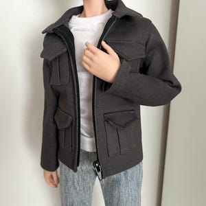 Könnte beinhalten: Eine dunkelgraue Jacke mit schwarzem Reißverschluss und vier Vordertaschen. Die Jacke wird über einem weißen T-Shirt getragen und mit hellblauen Jeans kombiniert. Die Jacke hat einen Kragen und ist für eine Puppe konzipiert.