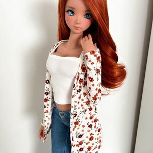 May include: A doll with long, auburn hair and blue eyes, dressed in a white ribbed vest top, a floral print cardigan, and distressed blue jeans. The cardigan has a pattern of red and brown flowers.