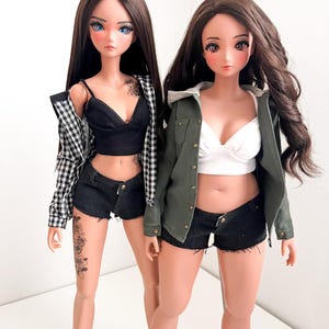 Könnte beinhalten: Zwei Puppen tragen schwarze Jeans-Shorts, eine mit einem schwarz-weiß karierten Hemd und die andere mit einer grünen Jacke. Beide Puppen haben langes braunes Haar und tragen schwarze BHs.