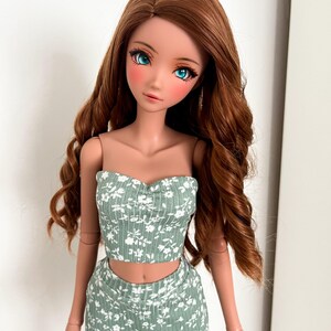 May include: A ball-jointed doll with long, wavy auburn hair and bright blue eyes. The doll wears a sage green, ribbed two-piece set consisting of a strapless tube top and a matching mini skirt, both featuring a small white floral pattern.