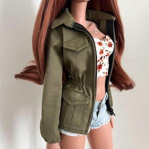 Könnte beinhalten: Eine Puppe trägt eine olivgrüne Jacke mit schwarzem Reißverschluss, ein weißes Oberteil mit Blumenmuster und Jeansshorts. Die Puppe hat lange, rotbraune Haare und posiert vor einem schlichten Hintergrund. Die Jacke hat zwei Vordertaschen und einen Kordelzug in der Taille.