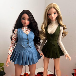 Puede incluir: Dos muñecas con atuendos diferentes. La muñeca de la izquierda lleva una falda y un chaleco de mezclilla azul con una camisa de estampado floral debajo. La muñeca de la derecha lleva una falda y un chaleco verdes con una camisa blanca de manga larga debajo.