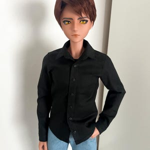 May include: A doll with brown hair and yellow eyes wearing a black button-down shirt and light blue jeans. The doll has a serious expression and is posed with one hand in a pocket. The shirt is buttoned up and has a collar.