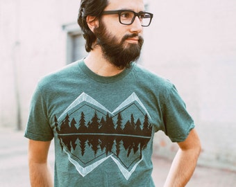 May include: A heathered green short-sleeve t-shirt featuring a graphic print of a black forest silhouette reflected within two white outlined diamond shapes. The design is centered on the chest of the crew neck garment.