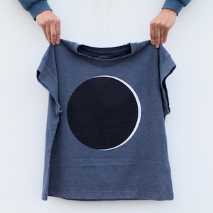 May include: A dark gray t-shirt featuring a large black circle with a white crescent design. The shirt is held up against a white background, showcasing the graphic print. The tee has short sleeves and a crew neck.