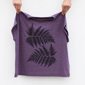 May include: A purple t-shirt featuring a black fern leaf design. The graphic shows two mirrored fern leaves. The shirt is held up by two hands against a white backdrop. The shirt is made of a soft material.