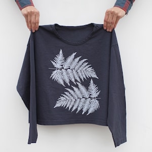 May include: A navy blue long-sleeved shirt featuring a light blue fern design. The fern design is composed of two large fern fronds with detailed leaf patterns. The shirt is held up against a white background.