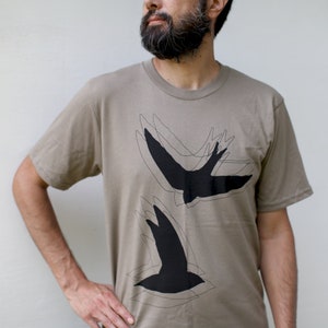 A close up chest shot of a light brown unisex t-shirt with 2 flying blackbirds on the front.