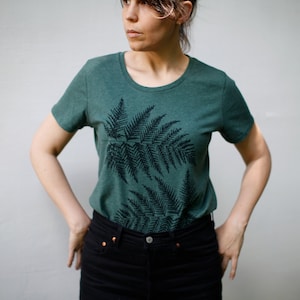 Puede incluir: Una camiseta verde azulada con un estampado de helecho negro. El dise&ntilde;o del helecho est&aacute; centrado en la parte delantera de la camiseta.