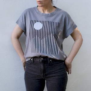 May include: A gray boxy top with black and white graphic print of a moon over snowy mountains. The shirt has cap sleeves and a round neck.