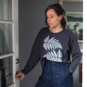 May include: A dark gray long-sleeve shirt featuring a white fern leaf design on the front. The shirt is paired with high-waisted, dark blue pants. The person is standing near a white door.