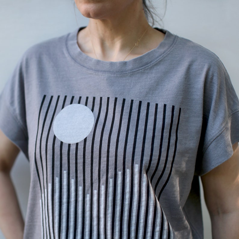 May include: A gray t-shirt with a black and white graphic design of vertical lines and a white circle.