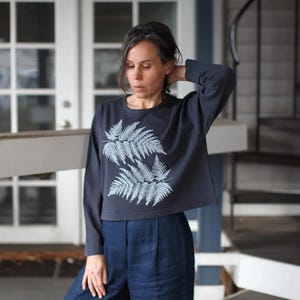May include: A navy blue long-sleeved shirt with a cropped hem, featuring a light blue fern design on the front. The shirt is paired with high-waisted, dark blue pants. The outfit is casual and suitable for everyday wear.