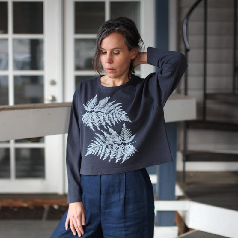 May include: A navy blue long-sleeved shirt with a cropped hem, featuring a light blue fern design on the front. The shirt is paired with high-waisted, dark blue pants. The outfit is casual and suitable for everyday wear.