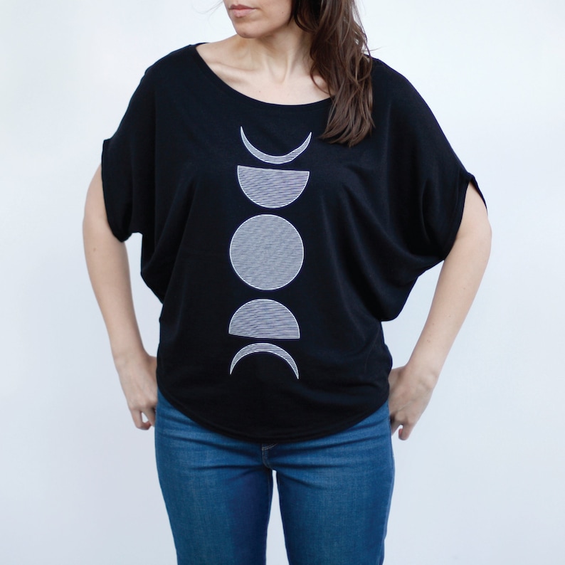 May include: A black t-shirt with a white graphic of a moon in different phases. The graphic is centered on the shirt and is a simple, modern design.