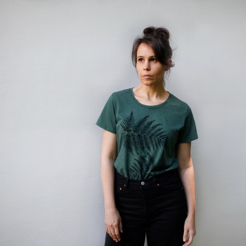 Puede incluir: Una mujer con una camiseta verde oscuro con estampado de helecho y pantalones vaqueros negros.