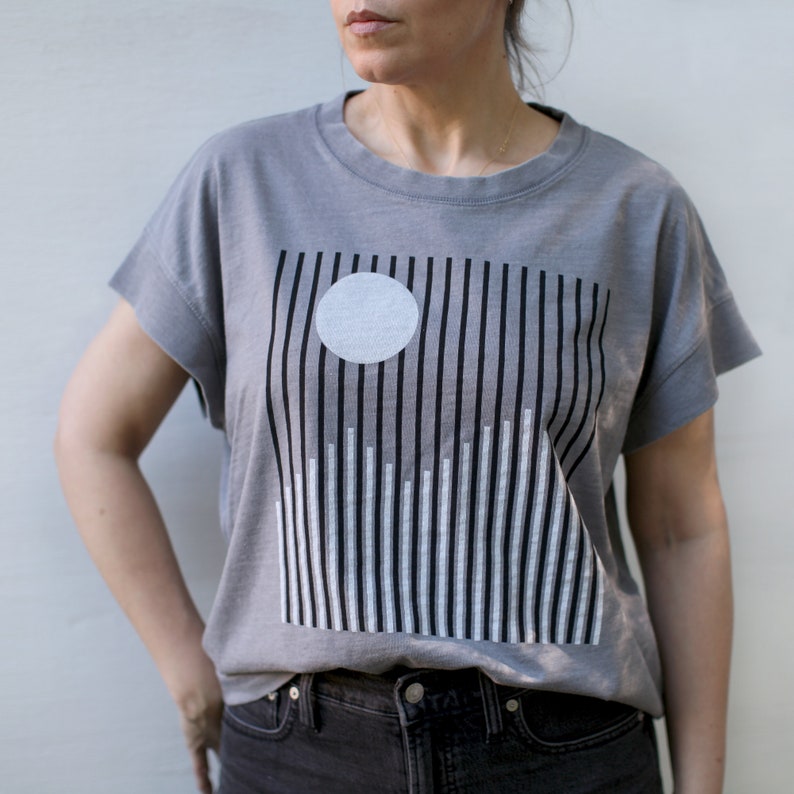 May include: A gray t-shirt with a graphic print of a white circle and black and white vertical lines.