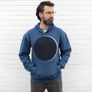 May include: A blue hoodie with a large black circle design and a white outline. The hoodie has a kangaroo pocket and a hood. The person in the image is wearing glasses and has a beard. The background is a white brick wall.